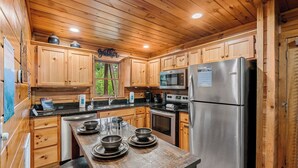 A wooden cabin kitchen with wooden cabinets, a stainless steel refrigerator, a microwave, and a wooden dining table with bowls and glasses. - Come getaway to a cabin in the Moonlit Pines (SEVIERVILLE)