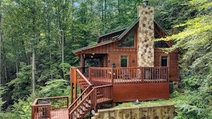 Une cabane dotée d’une cheminée en pierre, d’une terrasse en bois et d’un mur de soutènement en pierre.