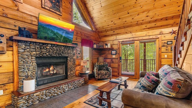 L’intérieur chaleureux d’un chalet en bois, avec une cheminée en pierre, un canapé confortable et une grande fenêtre donnant sur une forêt.