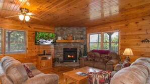Un intérieur de chalet chaleureux avec des murs en bois, une cheminée en pierre, un téléviseur et des canapés en cuir.