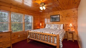 Une chambre à coucher dans une cabane en bois, avec un lit, deux tables de chevet, un ventilateur de plafond et de grandes fenêtres.