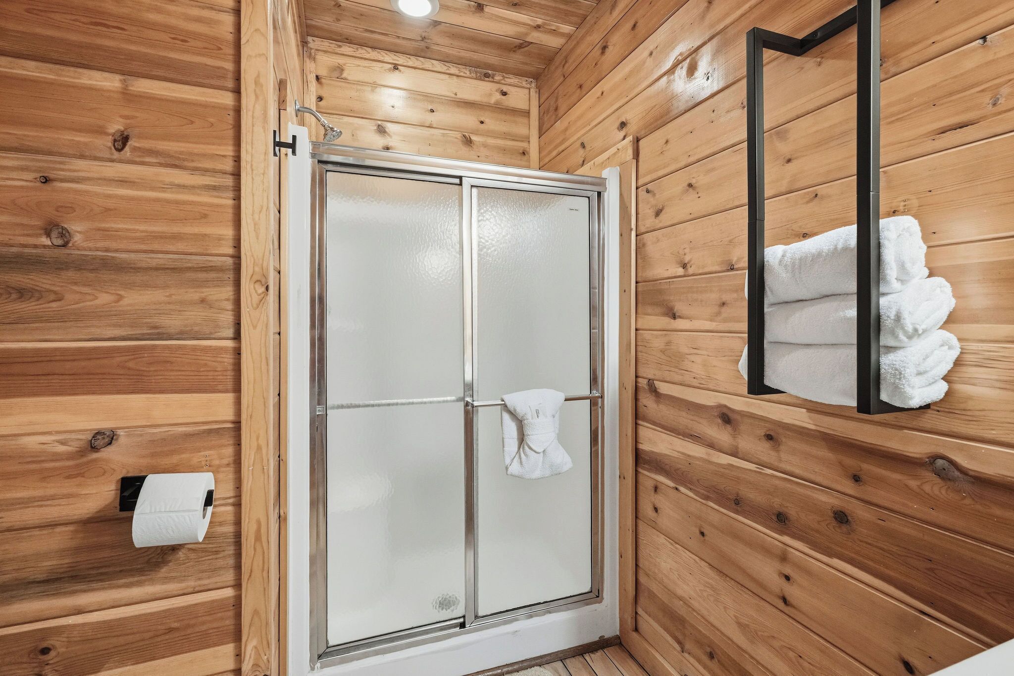 A wooden-paneled bathroom with a glass shower door, a towel rack, and a toilet paper holder.