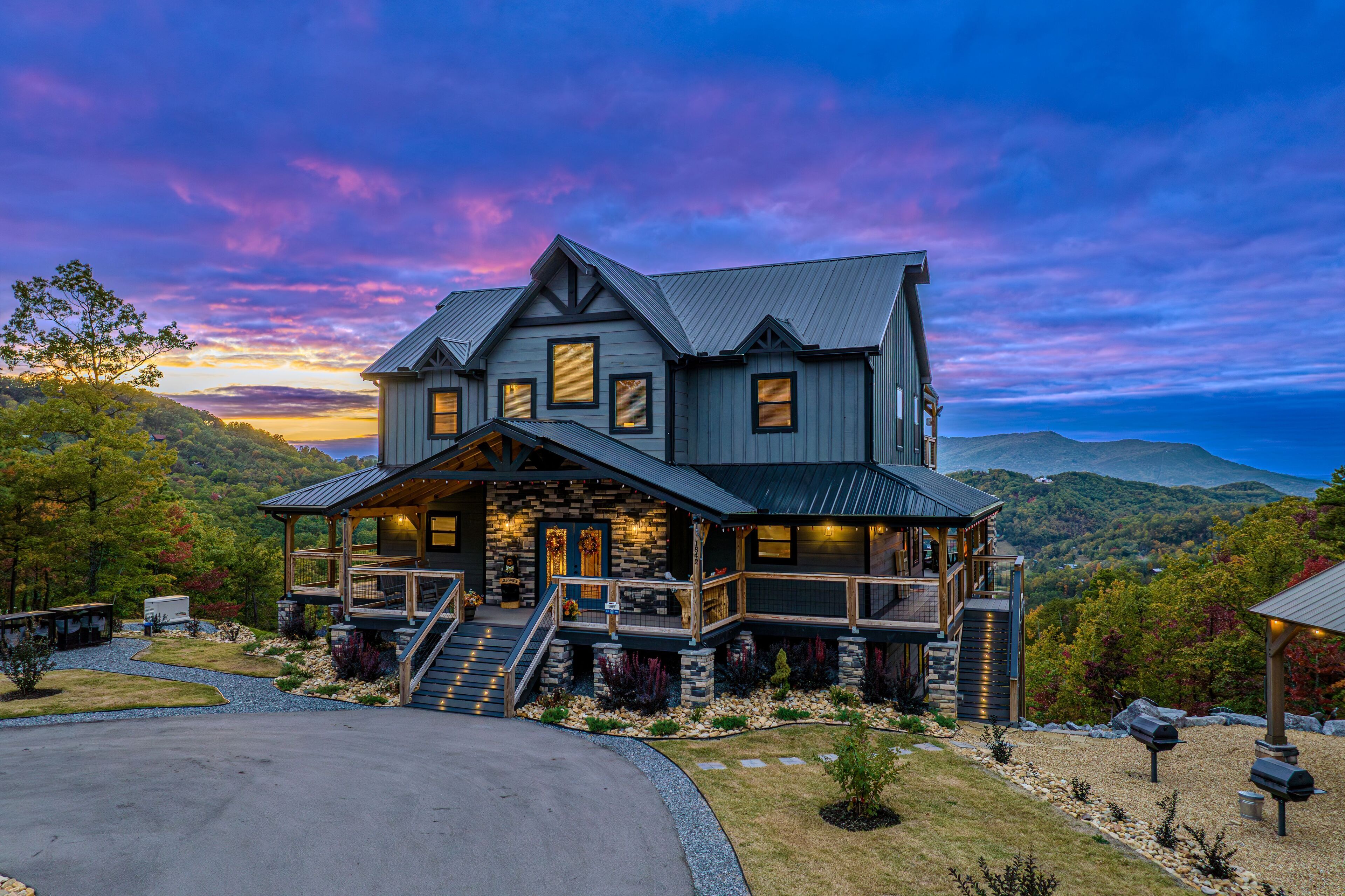 A large, multi-story house with a prominent front porch, stone accents, and a scenic mountain backdrop.