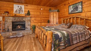 A log cabin interior with a stone fireplace, a bed with a plaid blanket, and a 'welcome' sign.