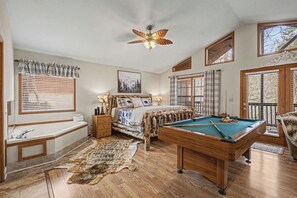 Une pièce avec une table de billard, un lit doté d’une tête de lit sur le thème de la forêt, un ventilateur de plafond et de grandes fenêtres équipées de stores.
