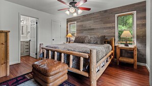 A bedroom with a wooden bed frame, a ceiling fan, a nightstand with a lamp, and a view of the outdoors through a window. - Mountain Views-Hot Tub-Fireplace-Close to Dollywood (SEVIERVILLE)