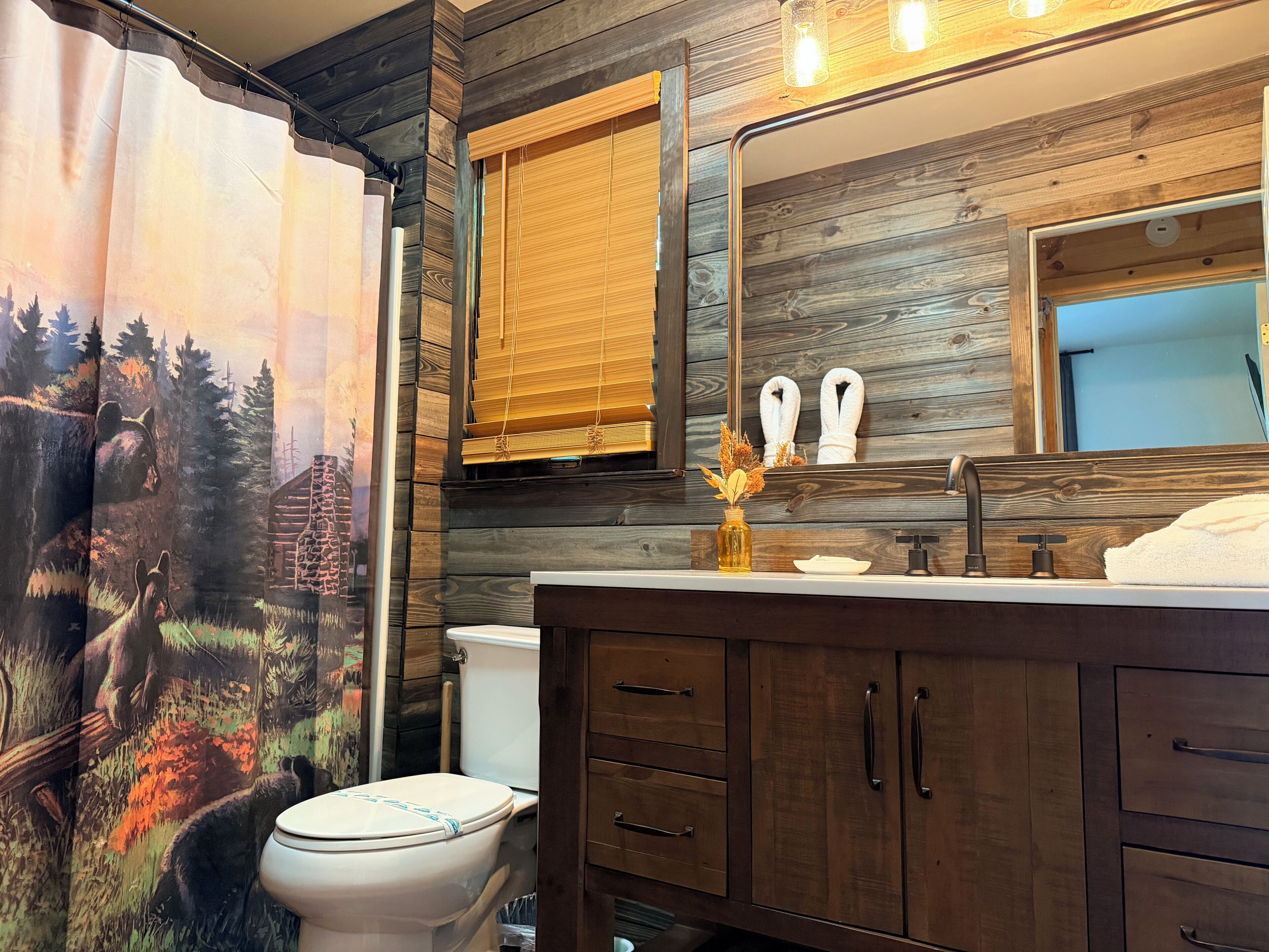 A bathroom with a wooden wall, a shower curtain with a forest scene, a toilet, a sink with a mirror, and a towel rack.