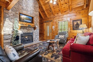 A log cabin interior with a stone fireplace, leather sofas, a wooden coffee table, and a television mounted above the fireplace.