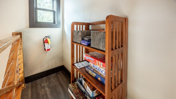Een hoek van een kamer met een houten boekenkast vol spelletjes, een brandblusser aan de muur en een raam met uitzicht op bomen.