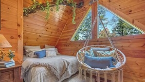 A cozy wooden cabin bedroom with a bed, a hanging chair, and a window with a view of trees. - Smoky Mountain Treehouse with hot tub and dog-friendly (GATLINBURG)