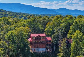 A two-story wooden cabin with a red roof, surrounded by dense forest and mountains in the background. - Surrounded By Mountain Beauty (GATLINBURG)