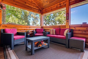A log cabin interior with wicker furniture, a glass-top coffee table, and a view of trees outside. - Surrounded By Mountain Beauty (GATLINBURG)