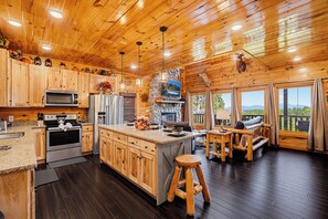 A cabin interior with wooden walls, a kitchen with modern appliances, a stone fireplace, and a deck with outdoor seating. - Surrounded By Mountain Beauty (GATLINBURG)