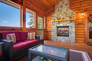 A log cabin interior with a stone fireplace, wicker furniture, and a clear view outside. - Surrounded By Mountain Beauty (GATLINBURG)