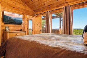 A wooden cabin room with a bed, a television mounted on the wall, and a view of the outdoors through a window.
