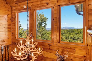 A wooden cabin interior with a chandelier and two windows offering a view of a forest and mountains. - Surrounded By Mountain Beauty (GATLINBURG)