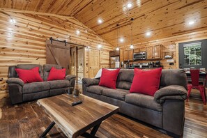 A cozy cabin interior with a living area featuring two sofas with red cushions, a wooden coffee table, and a kitchen area with modern appliances.
