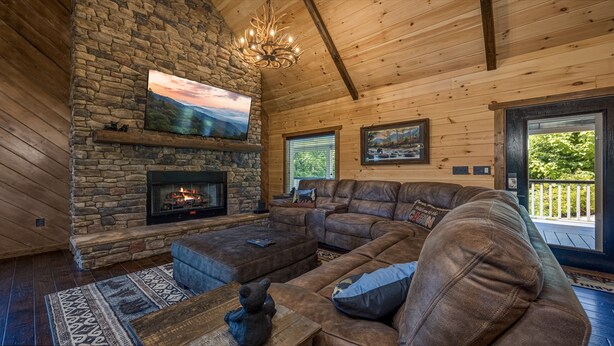 A cozy cabin interior with a stone fireplace, leather sectional sofa, and a large TV mounted on the wall. - If Every View Could Be This Perfect (GATLINBURG)