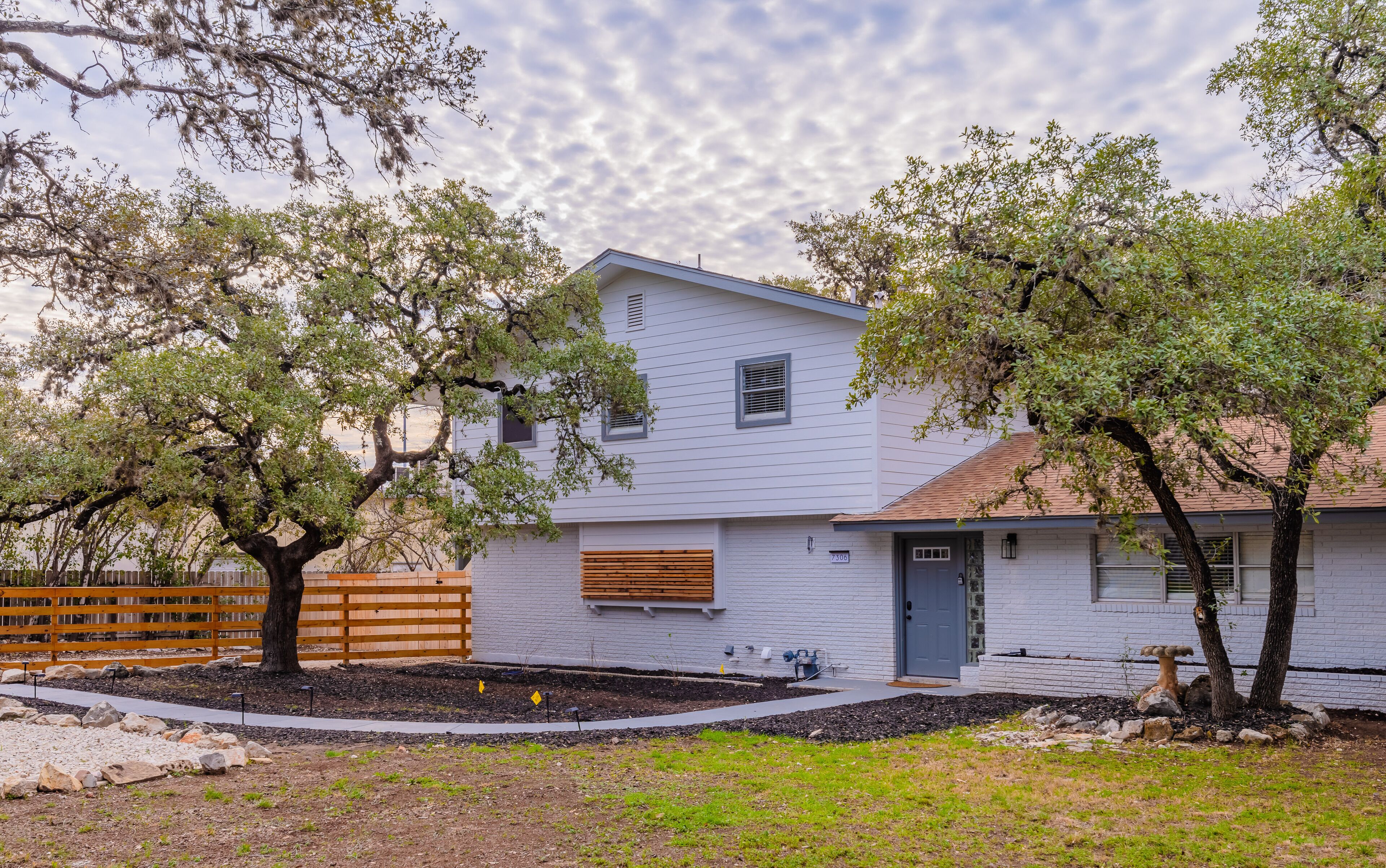 A two-story house with a white exterior, a front garage, and a small front yard with a wooden fence and trees.
