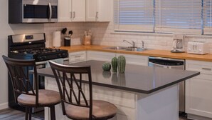 A modern kitchen with white cabinets, a stainless steel microwave, a gas stove, and a dishwasher. There are two wooden chairs with cushioned seats and a small island with two decorative vases. - Hot Tub, Unique Tree Deck & Game Room near Six Flags Fiesta Texas (San Antonio)
