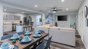 A modern kitchen with white cabinets, a dining area with a wooden table set for four, and a living area with a sofa and a TV mounted on the wall. - Hot Tub, Unique Tree Deck & Game Room near Six Flags Fiesta Texas (San Antonio)