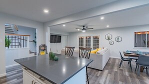 A modern kitchen with a central island, a dining area with a sofa and chairs, and a staircase leading to an upper level. - Hot Tub, Unique Tree Deck & Game Room near Six Flags Fiesta Texas (San Antonio)