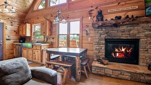 A cabin interior with a stone fireplace, wooden furniture, and a kitchen area. - Taking Hide And Go Seek To The Next Level (PIGEON FORGE)
