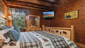 Dormitorio de cabaña de madera con una cama estampada con motivos de animales, un televisor montado en la pared y un letrero de advertencia sobre osos.