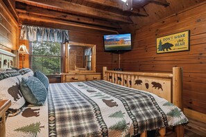 Dormitorio de cabaña de madera con una cama estampada con motivos de animales, un televisor montado en la pared y un letrero de advertencia sobre osos.