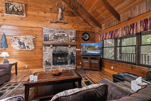 A cozy cabin interior with a stone fireplace, wooden walls, a television, and comfortable seating.