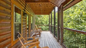Une terrasse en bois, agrémentée de quatre chaises et d’une table, également en bois, et entourée d’une rambarde en métal, offrant une vue sur une forêt verdoyante et luxuriante.