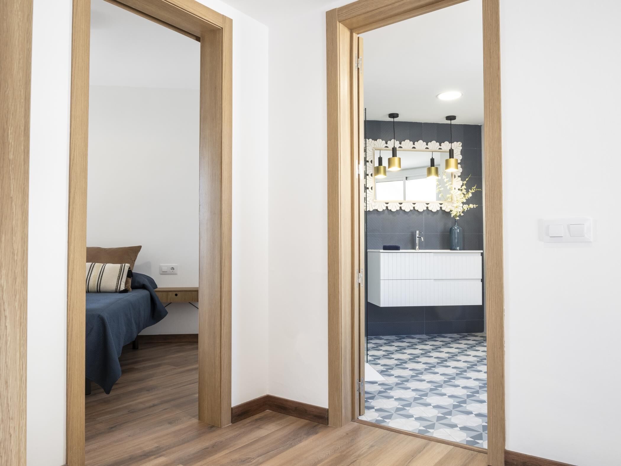 Un dormitorio moderno con cama, suelo de madera y baño con suelo de baldosas, lavamanos e iluminación colgante.