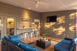 A modern outdoor living space with a ceiling fan, a bar area with stools, and a 'Happy Hour' sign. - Mid-Century Retreat with Private Pool near Airport, Pearl & Downtown (San Antonio)