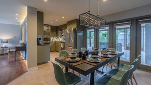 A modern kitchen with a dining area, featuring a central island, stainless steel appliances, and a large window providing a view of the outdoors. - Private Jetted POOL, Firepit & BBQ in a Stunning House By Airport (San Antonio)