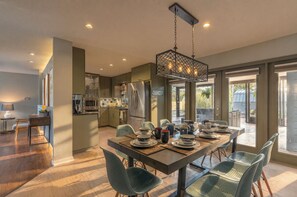 A modern kitchen with stainless steel appliances, a dining area with a long table and chairs, and a view of the outdoors through large sliding doors.