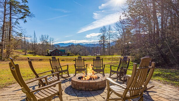 A fire pit with chairs arranged around it, set on a paved area with a view of mountains and a house in the background. - Firepit - Mountain Views - Car Show Friendly (GATLINBURG)
