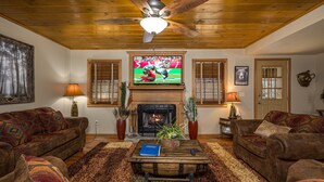 A cozy living room with a fireplace, two sofas, a coffee table with a book, and a television displaying a sports game. - Firepit - Mountain Views - Car Show Friendly (GATLINBURG)