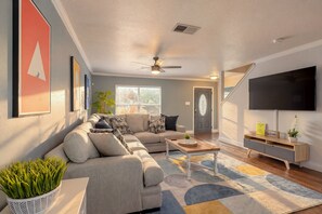 A living room with a sectional sofa, a coffee table, a flat-screen TV, and a staircase. - Private Hot Tub & Game Room near SeaWorld and Lackland AFB (San Antonio)