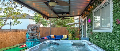 Un jacuzzi bajo un porche cubierto con ventilador de techo, cerca de una valla de madera y una pared verde con plantas trepadoras.