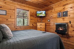 A wooden cabin room with a bed, a television, a fireplace, and a window with blinds.