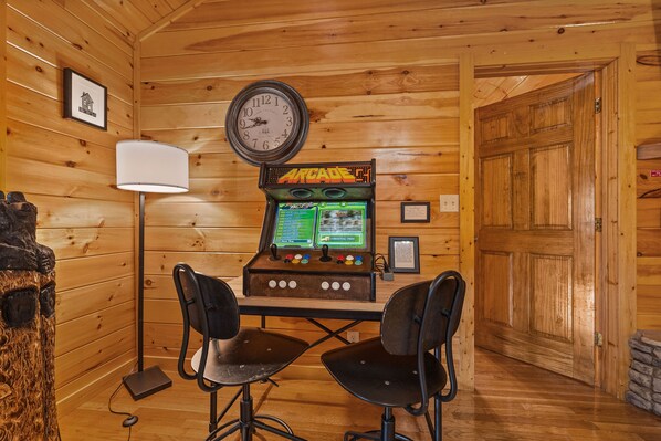 Ein Holzinterieur mit einer Vintage-Spielmaschine, zwei schwarzen Stühlen, einer Wanduhr und einem gerahmten Bild.