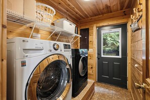 A laundry room with two washing machines, wooden walls, a door with a view of trees, and a shelf with laundry baskets.