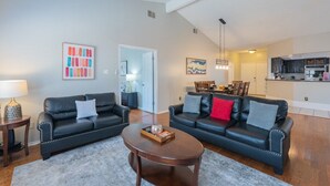 A modern living room with two black leather sofas, a wooden coffee table, and a gray rug. There is a kitchen visible in the background with a dining area.