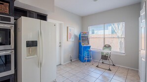 A kitchen with a white refrigerator, a blue arcade machine, and a baby high chair near a window.