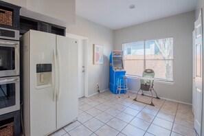 Une cuisine avec un réfrigérateur blanc, une borne d’arcade bleue et une chaise haute pour bébé près d’une fenêtre.