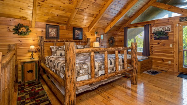 Une chambre dans un chalet en rondins, avec un lit en bois, une table de chevet, une lampe, un tableau encadré et une fenêtre avec des rideaux.