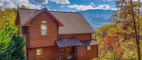 A wooden cabin with a porch, surrounded by autumn foliage and mountains in the background.