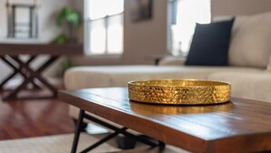 Een woonkamer met een houten salontafel, een gouden decoratieve schaal, een bank en een raam met natuurlijk licht.