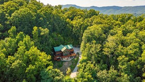 A large, two-story house surrounded by dense forest, with a visible driveway leading to the entrance.