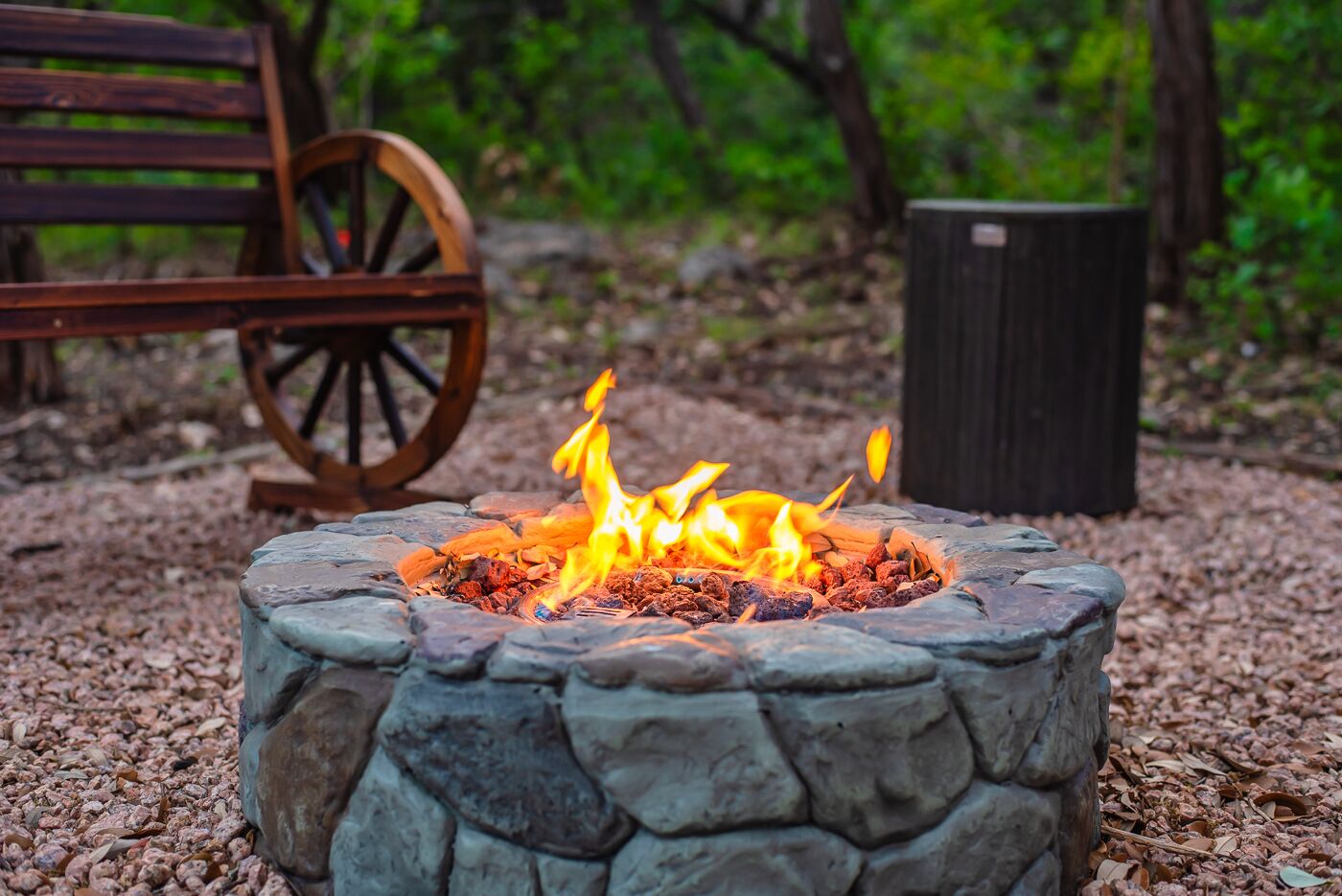 A stone fire pit with flames, surrounded by gravel, with a wooden bench and a wheelbarrow in the background.